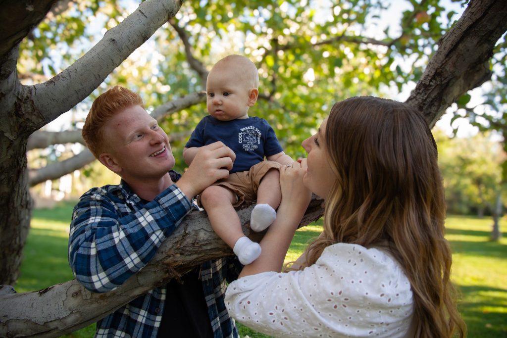 Family gathered around, admiring their son sitting in a tree, captured by Kendalyn Rosenlund, a professional family photographer